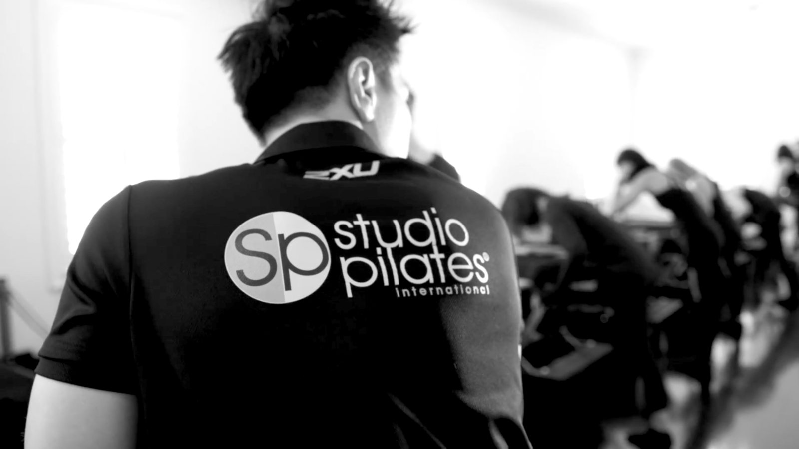 Person wearing a shirt with studio pilates international logo facing a group doing Pilates indoors.