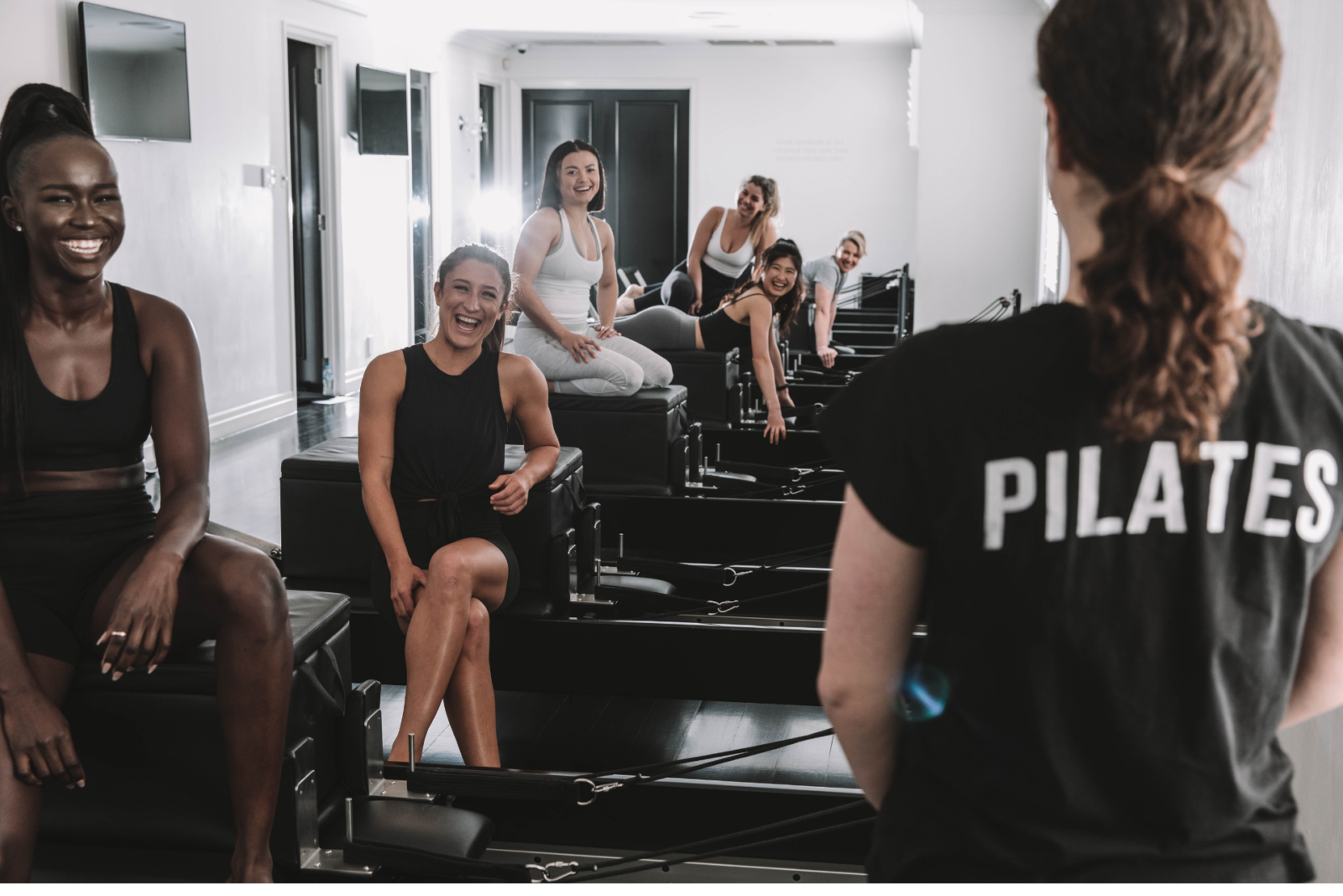 Women in a Pilates class use reformer machines while interacting with the instructor in a bright studio.