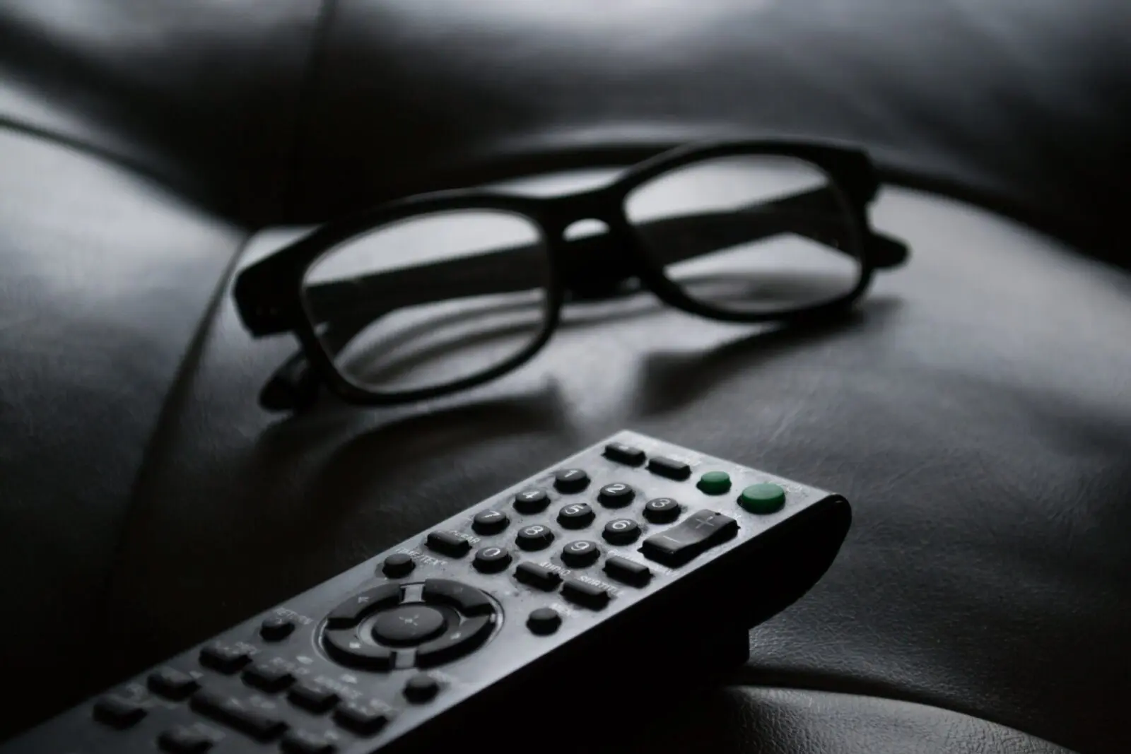 Black TV remote control and black-framed eyeglasses on a dark leather surface.