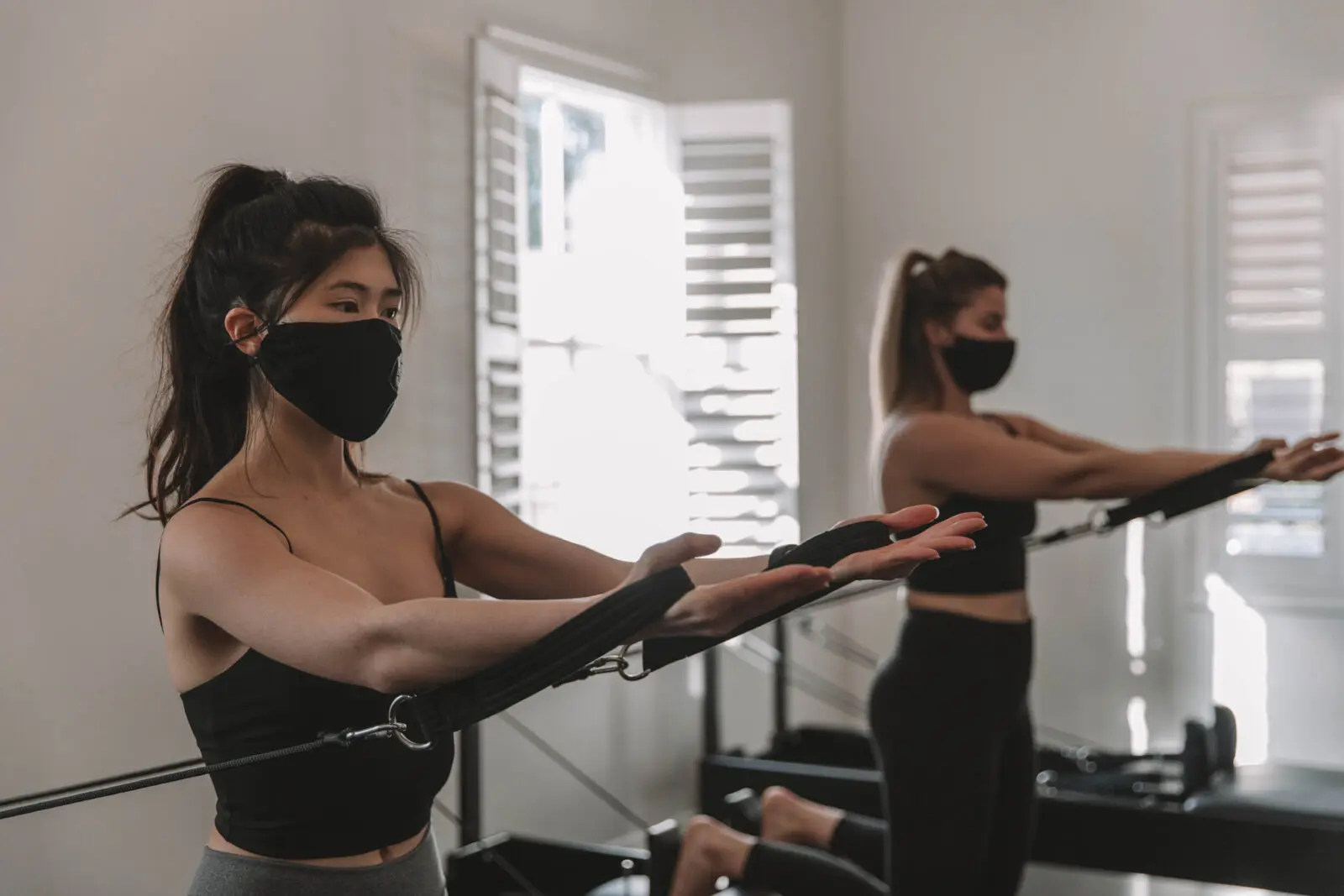 Two women in black masks and fitness wear exercising with resistance bands in bright room.