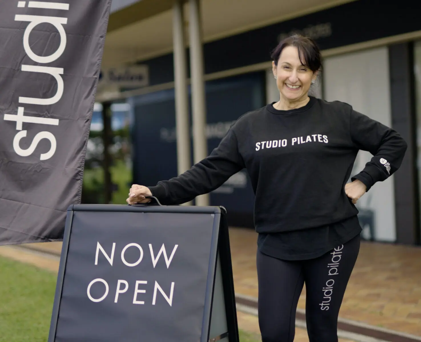 Woman in Studio Pilates attire stands outside near a now open sign at the studio entrance.