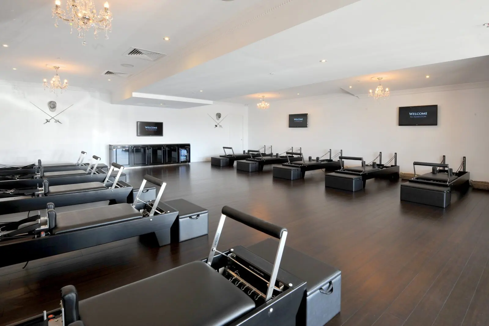 Fitness studio with black Pilates reformers, crystal chandeliers, flat-screen TVs, and crossed swords decor.