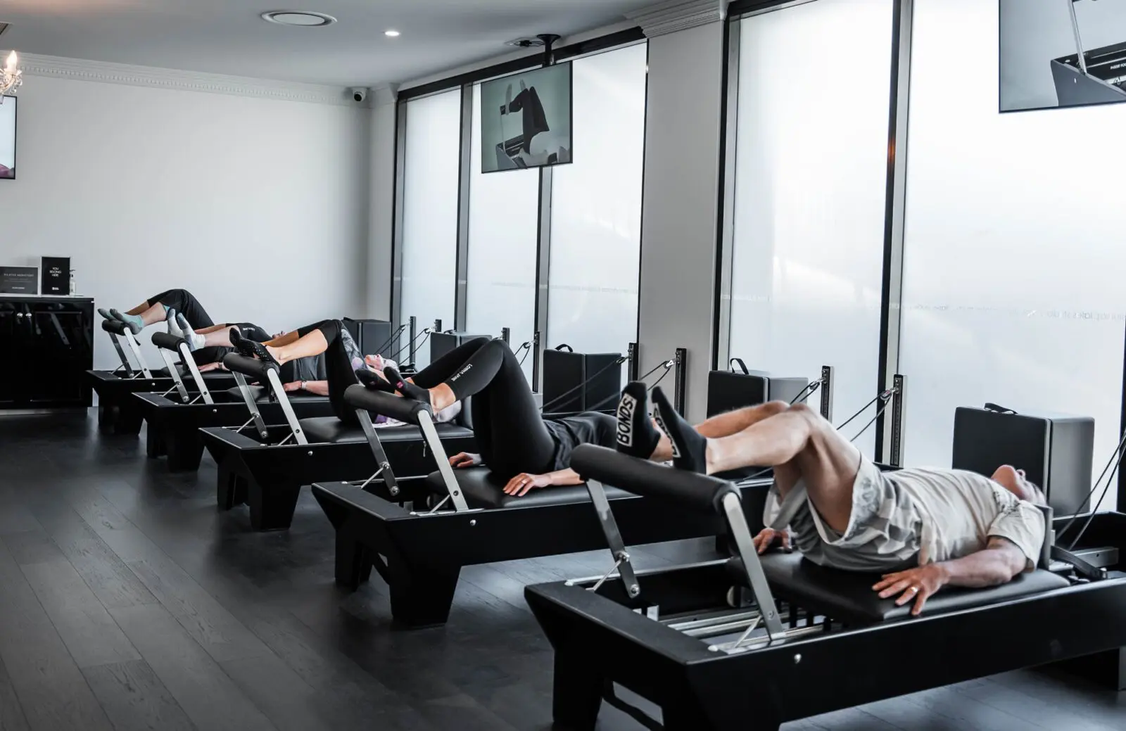 People exercising on Pilates reformer machines in a bright studio with large windows.