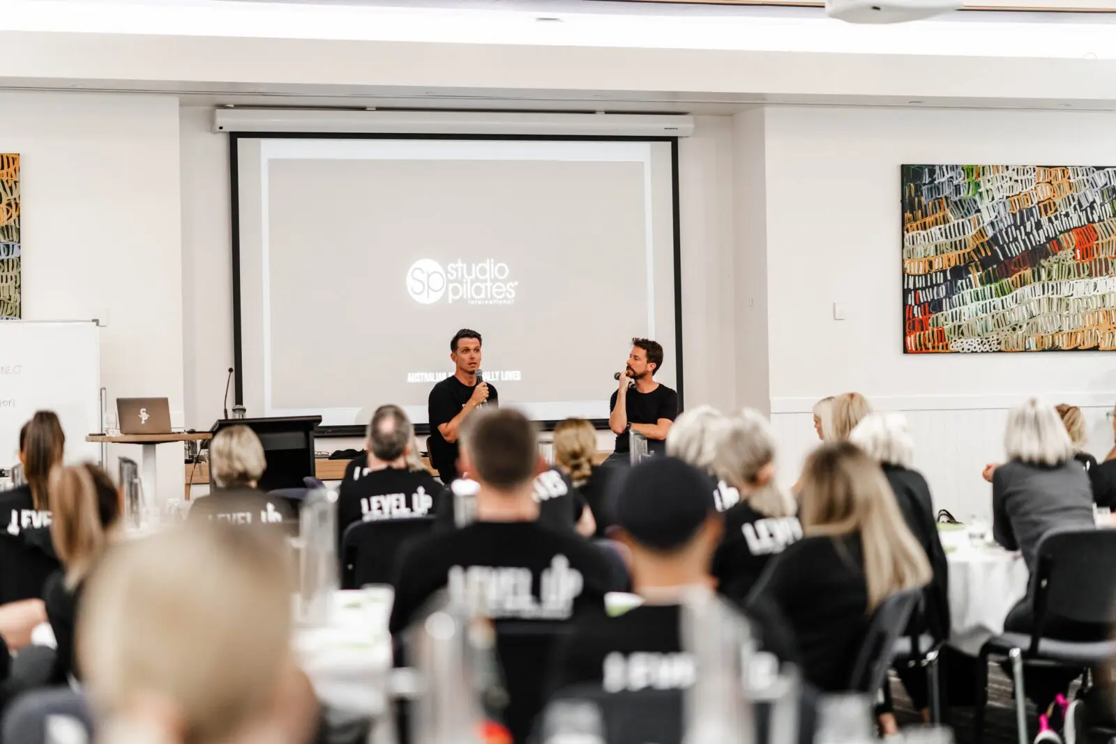 Audience in matching shirts listens to two speakers in front of a Studio Pilates International presentation screen.