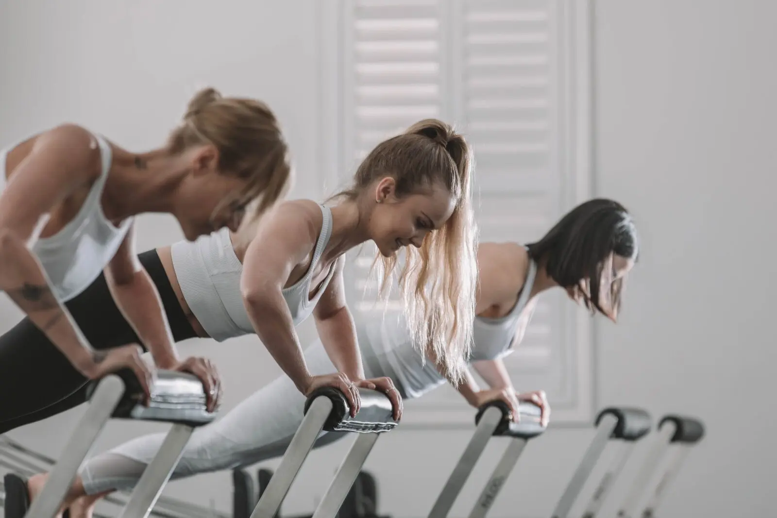 Three women performing inclined push-ups using fitness equipment in a bright workout studio.