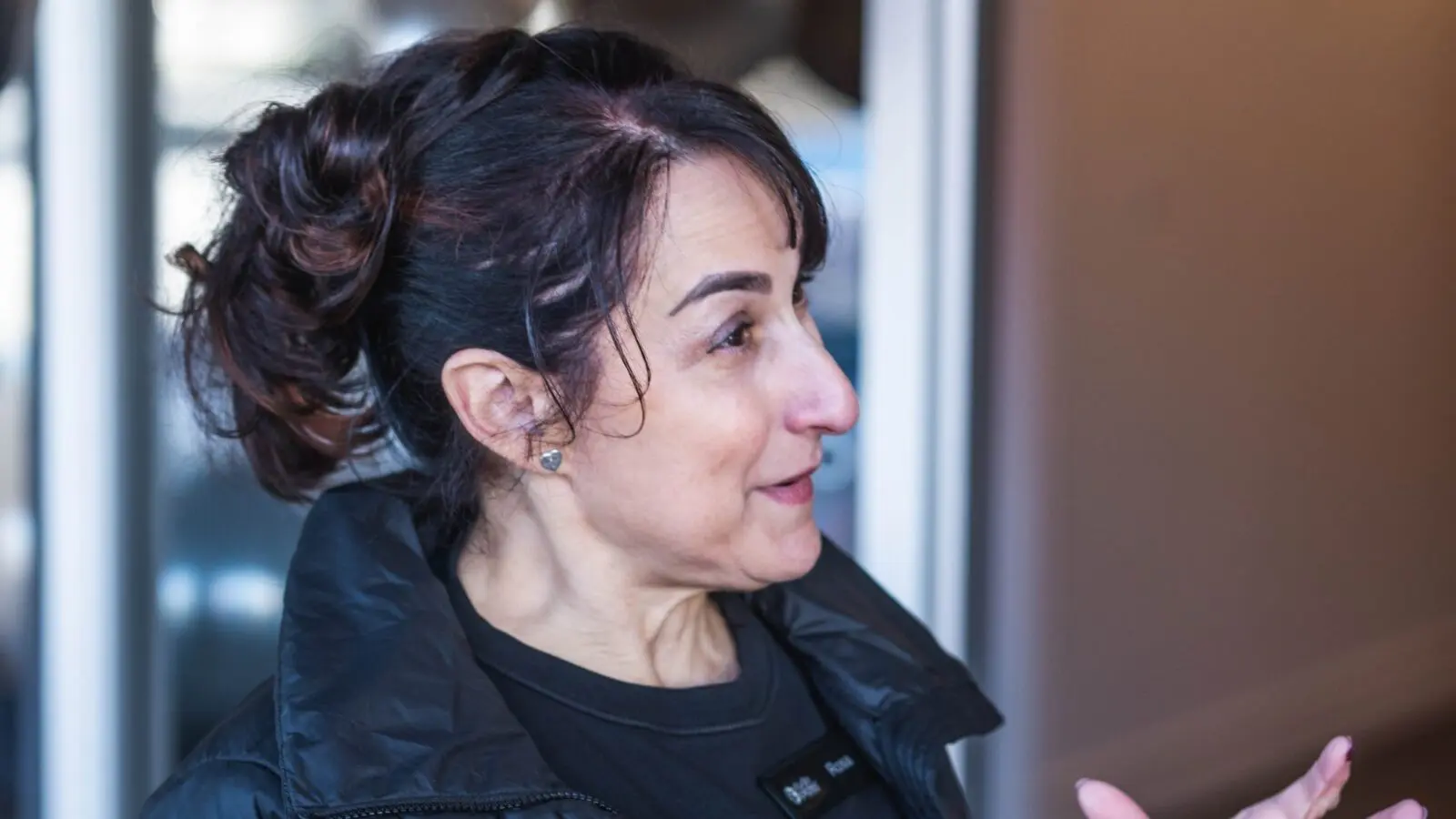 Profile view of a woman with dark hair in a bun wearing a black jacket indoors.