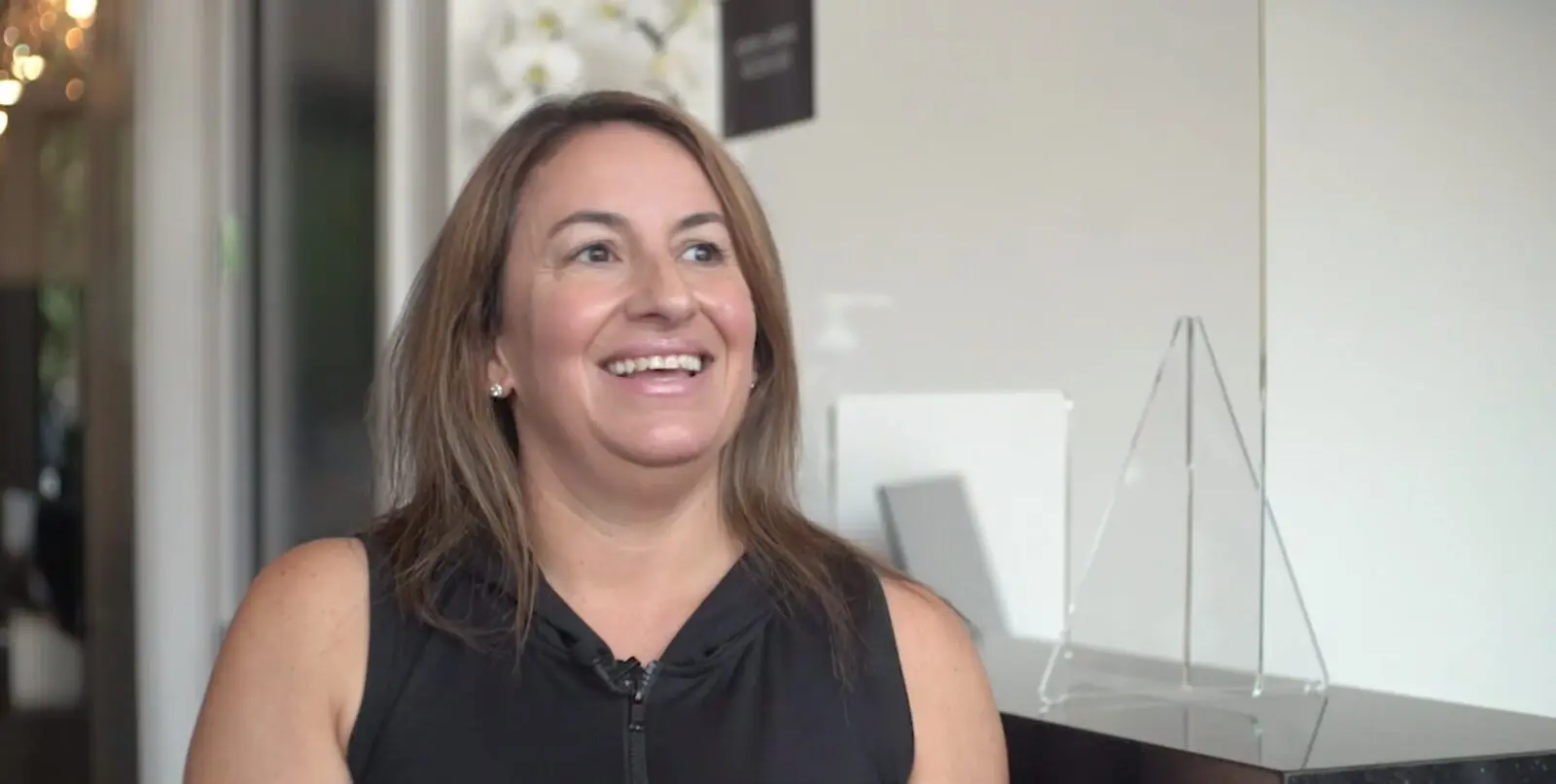 Woman with shoulder-length brown hair in sleeveless black top smiling indoors near clear triangular stand.