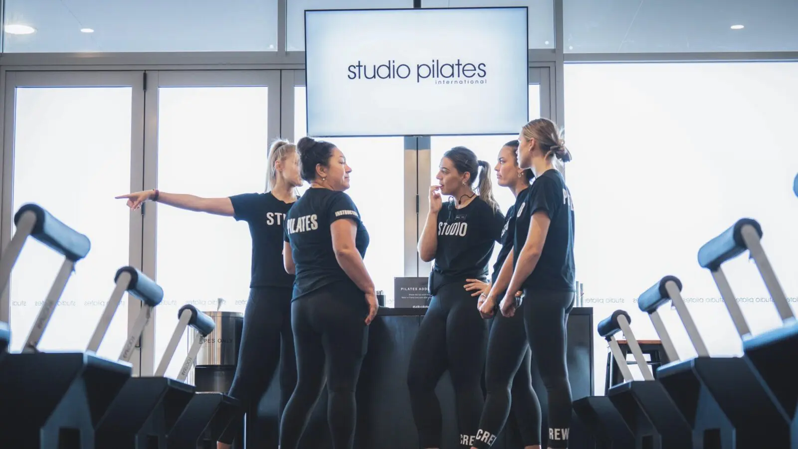 Five women in black workout clothes talking inside a Pilates studio with reformer machines.