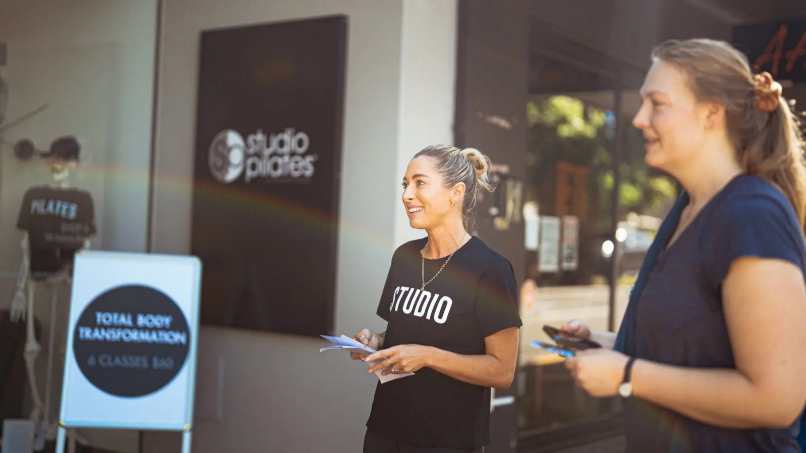 Two women stand outside a Pilates studio with a sign advertising six classes for $60.