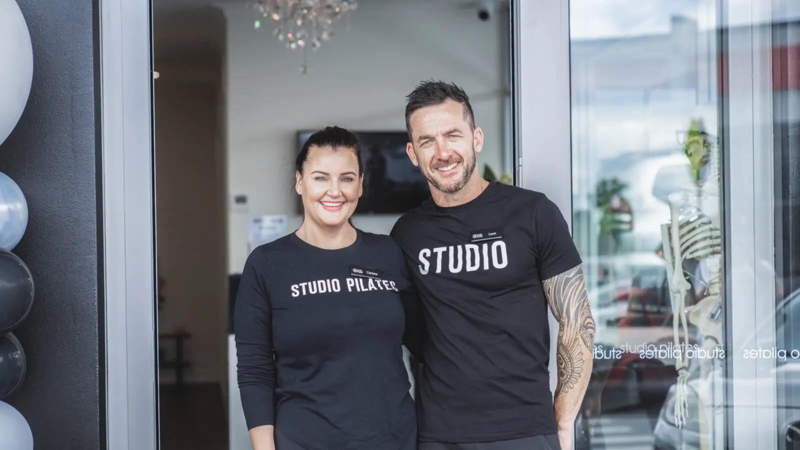 Two staff members wearing Studio Pilates shirts stand at a studio entrance with balloons and a skeleton model.