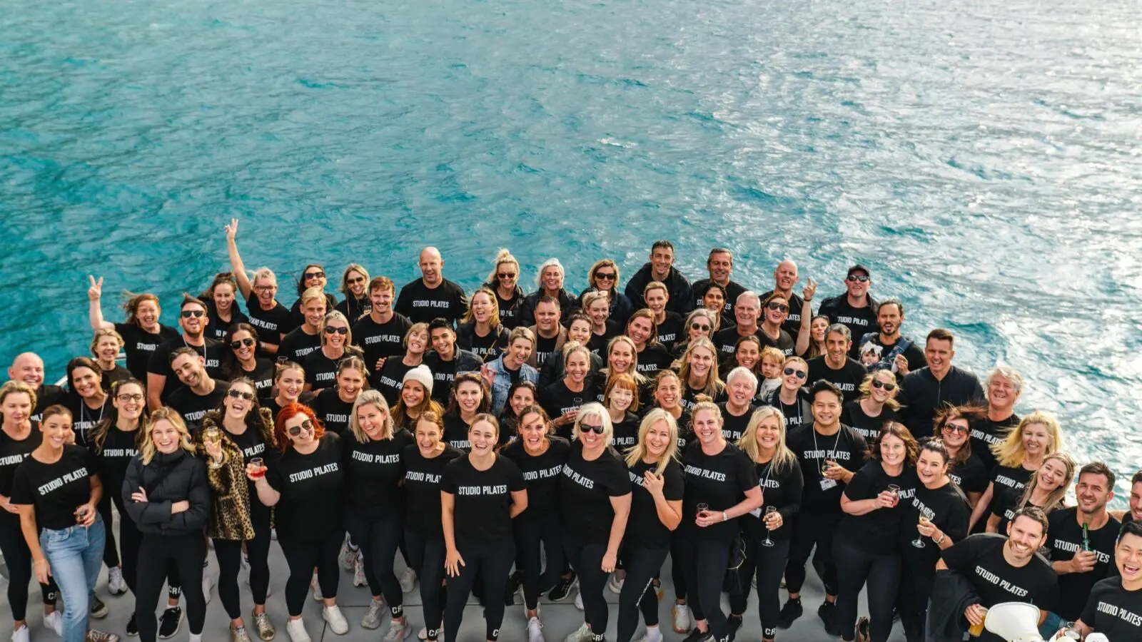 Group of people wearing matching Studio Pilates shirts posing on a deck by the water.