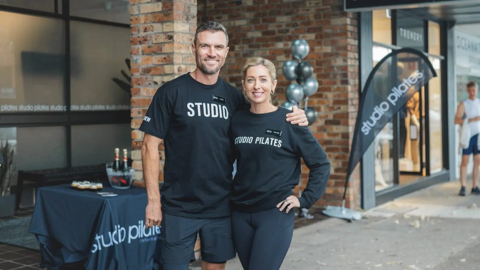 Two people in black Studio Pilates apparel stand outside a brick building near a branded table and flag.