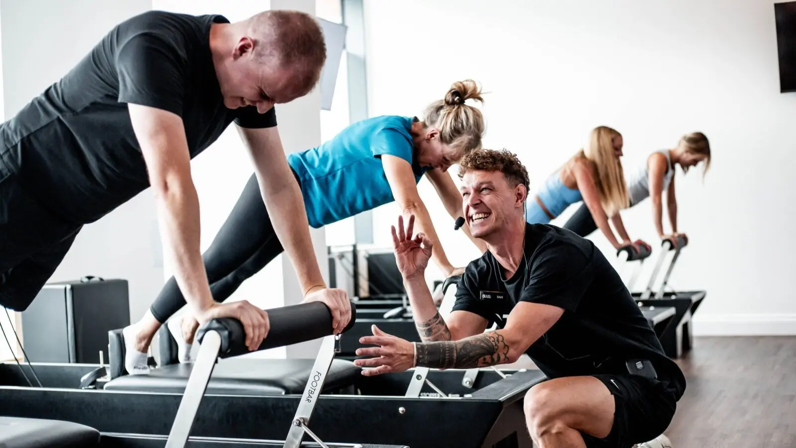 Fitness instructor coaching three people on reformer machines performing plank exercises in a gym.