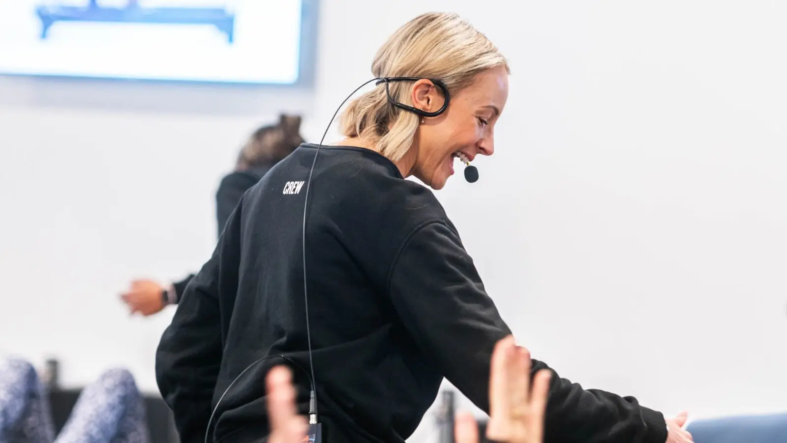 Person wearing a black crew sweatshirt and headset microphone interacting with raised hands.