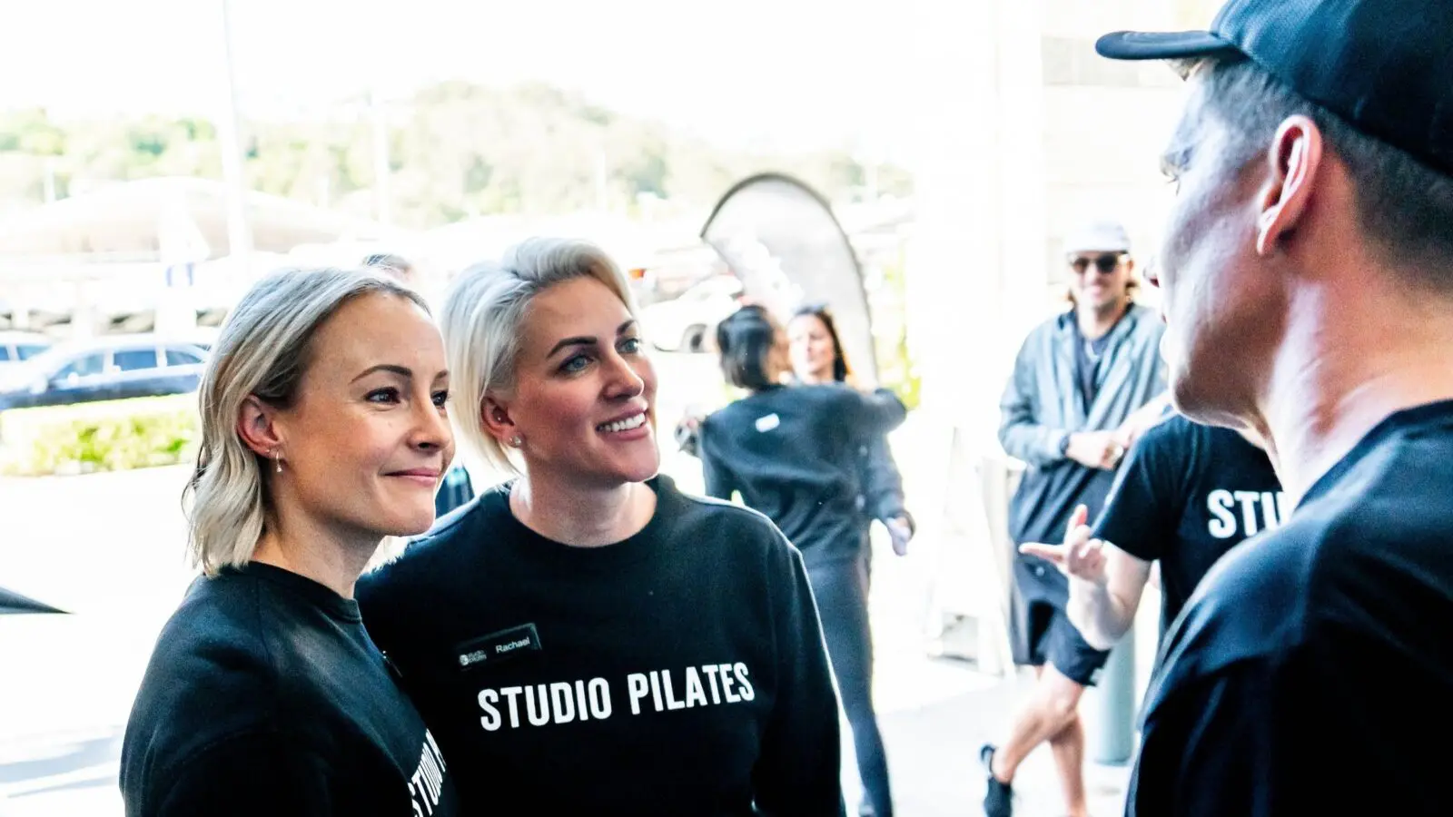 Two women in Studio Pilates shirts talking to a man outdoors with people and cars nearby.