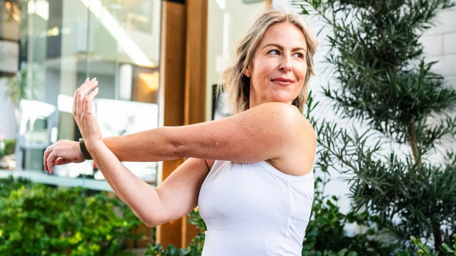 Woman in white tank top stretching her arm outdoors near plants and glass building.