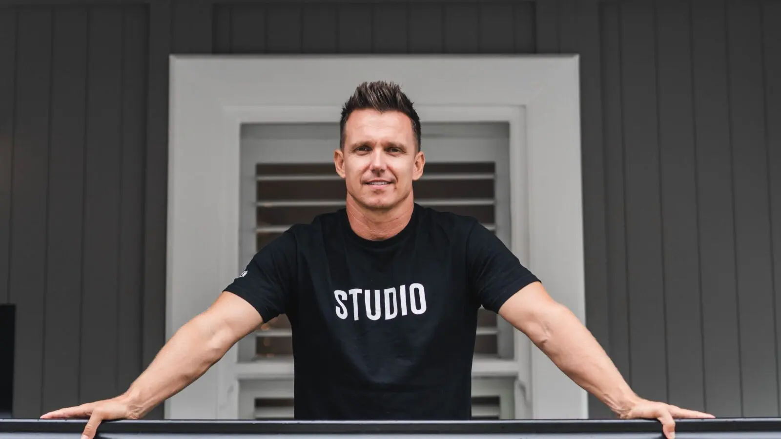 Man in black t-shirt with STUDIO text leaning on a railing in front of a window with shutters.