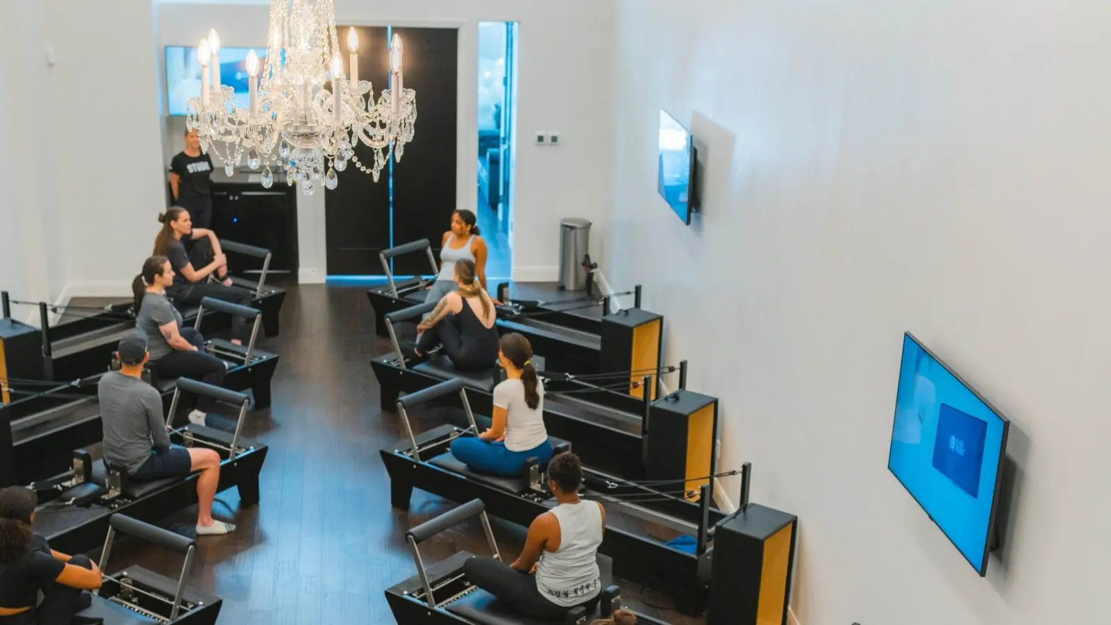 People seated on Pilates reformer machines in a spacious room with chandelier and wall-mounted TVs.
