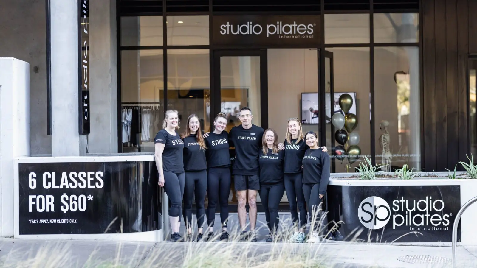Seven people in black attire stand outside Studio Pilates International with promotional signage visible.