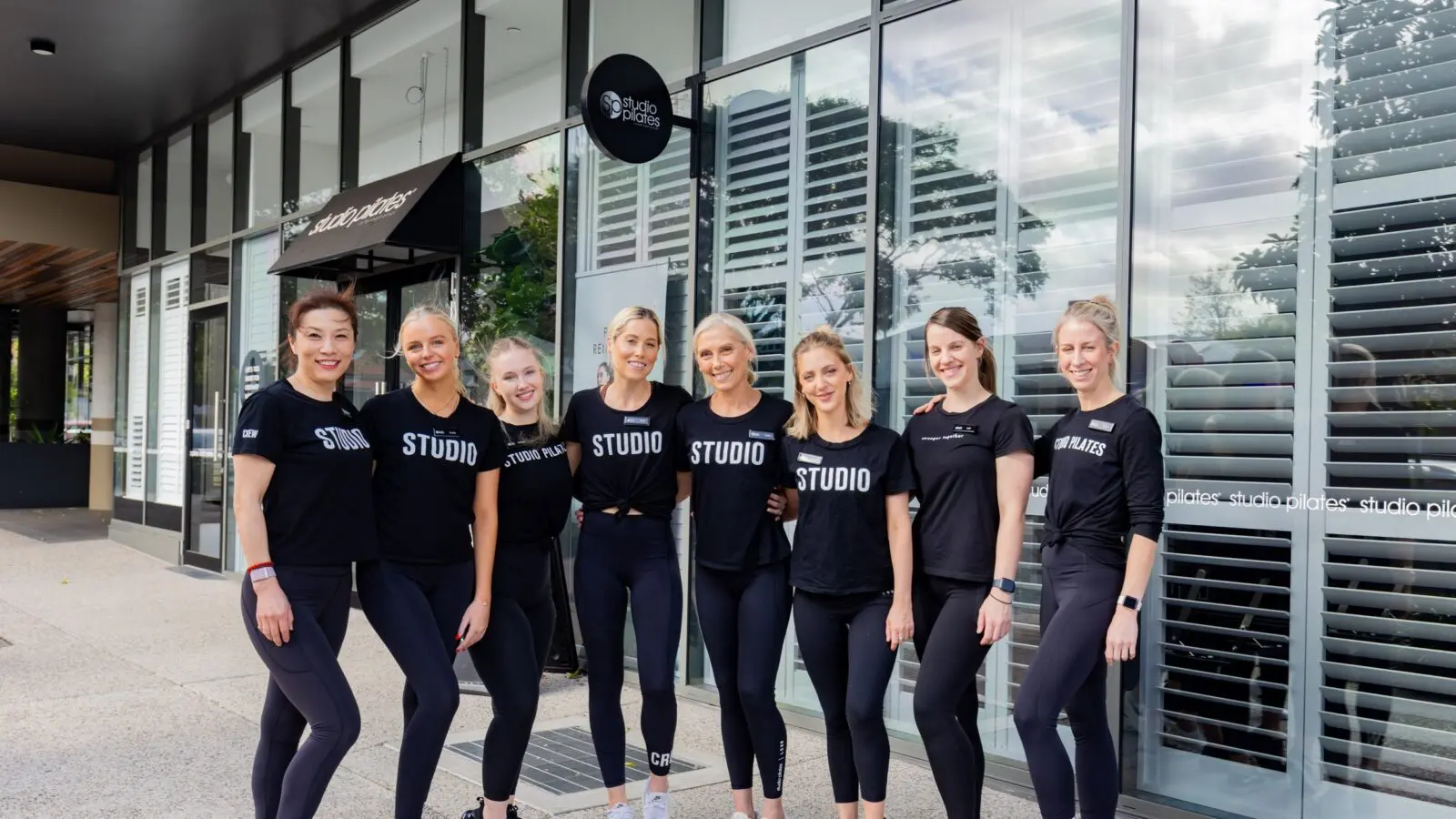 Nine women in black athletic wear stand outside Studio Pilates with glass windows and signage.