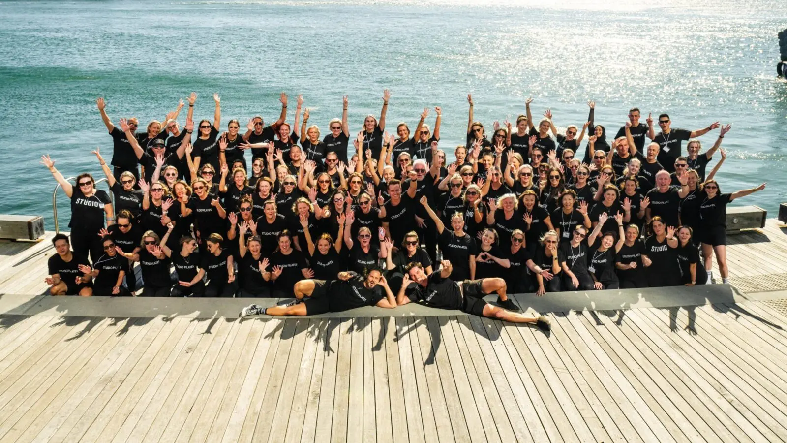Large group in black Studio Pilates shirts posing with raised hands on wooden dock by water.