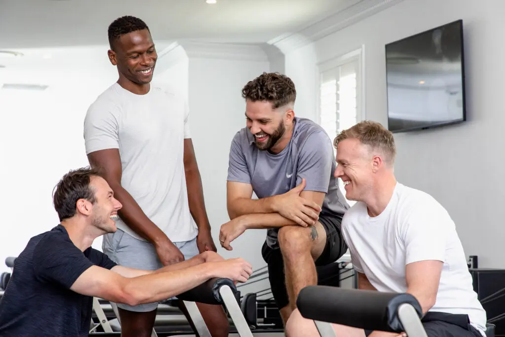 Four men in workout attire smiling and talking in a fitness room with exercise equipment.