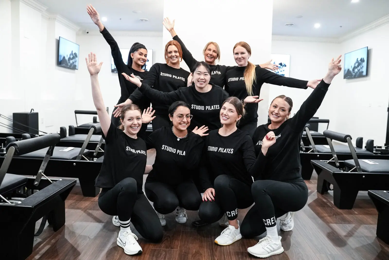 Nine women wearing Studio Pilates shirts posing with arms raised inside a Pilates studio with reformer machines.