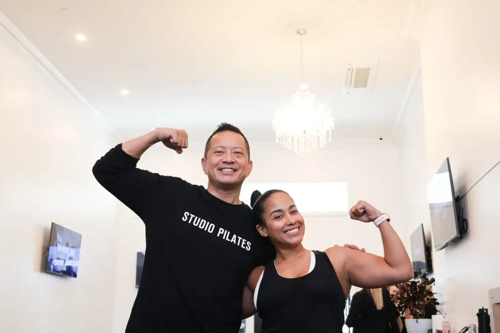 Two people flexing arm muscles inside a well-lit Pilates studio with screens and chandelier.