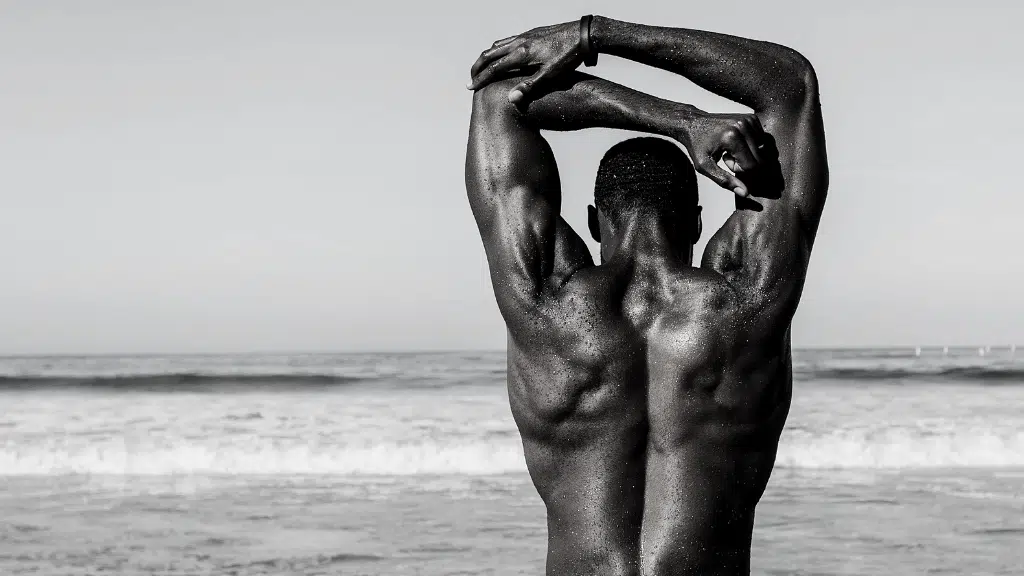 Muscular man stretching with arms raised at the beach facing calm ocean in black and white.