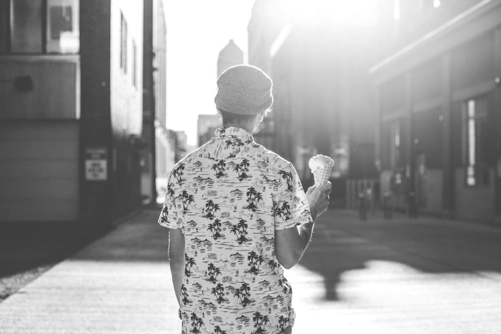 Person in a patterned short-sleeve shirt and beanie holding an ice cream cone on an urban street.