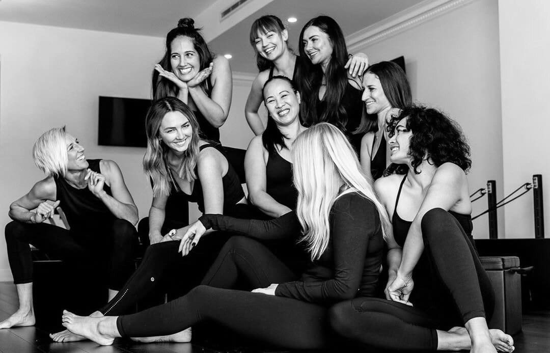 Nine women in athletic wear sitting and standing together indoors, smiling and interacting.