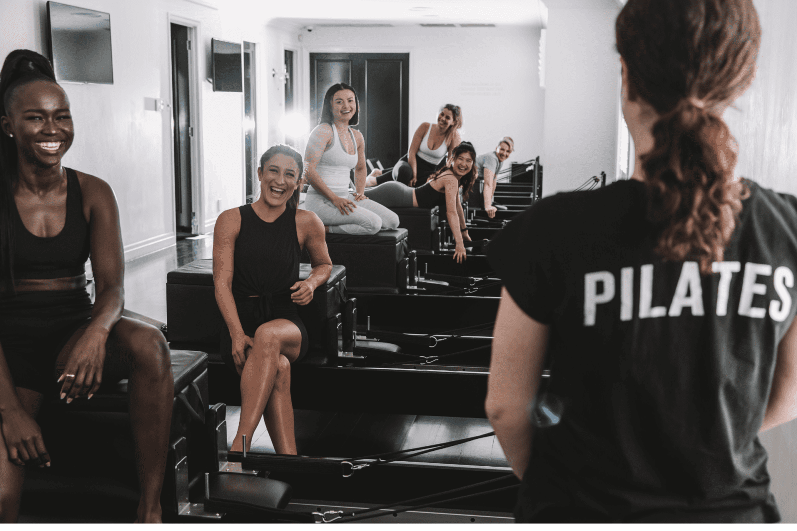Women in a Pilates class use reformer machines while interacting with the instructor in a bright studio.