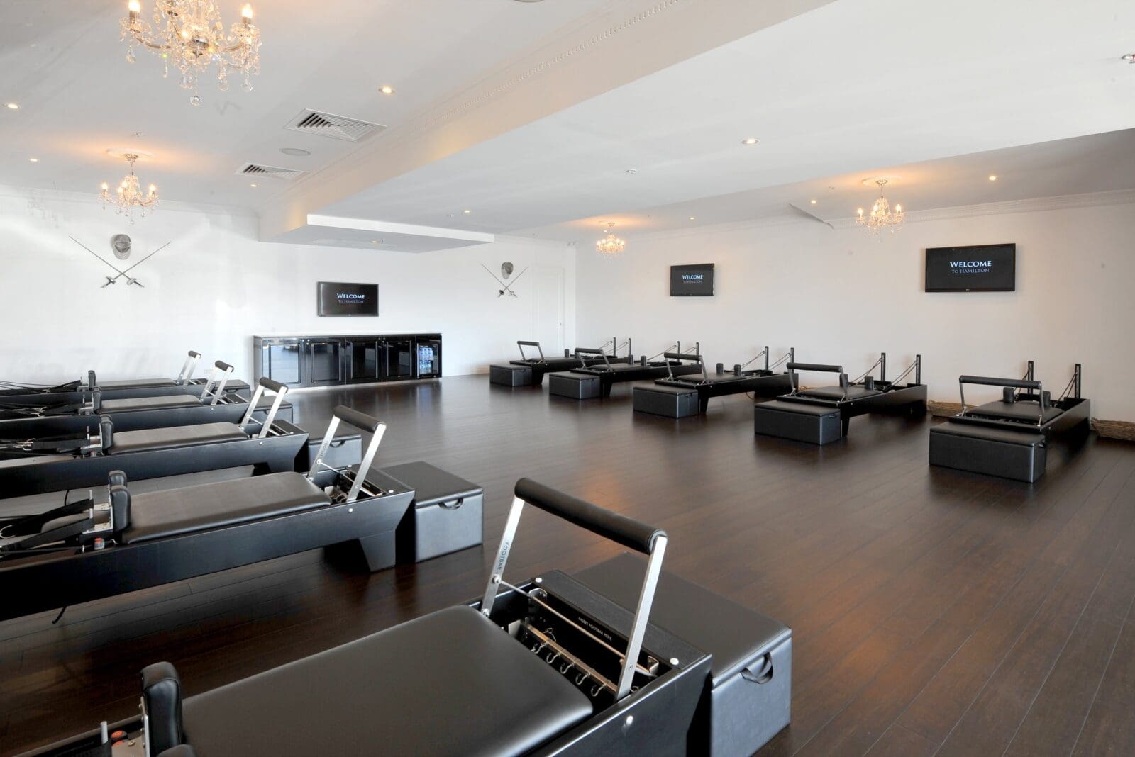 Fitness studio with black Pilates reformers, crystal chandeliers, flat-screen TVs, and crossed swords decor.