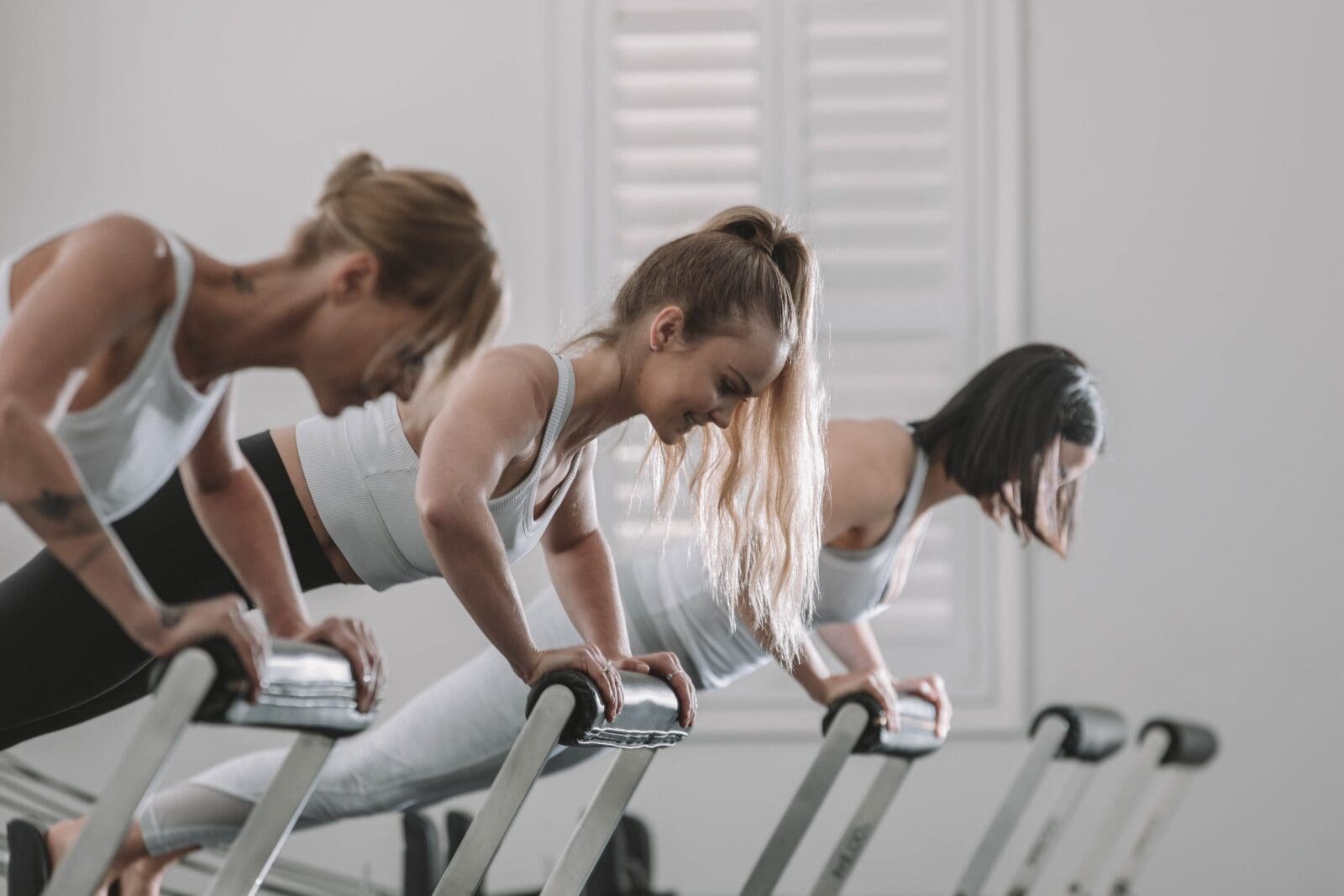 Three women performing inclined push-ups using fitness equipment in a bright workout studio.