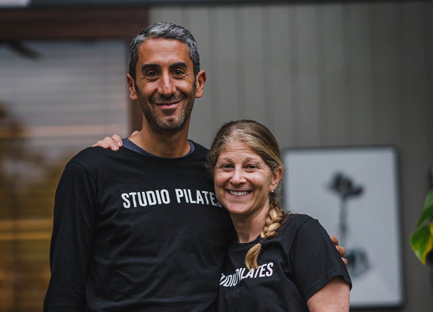 A man and woman smiling indoors wearing black Studio Pilates shirts with a blurred background.