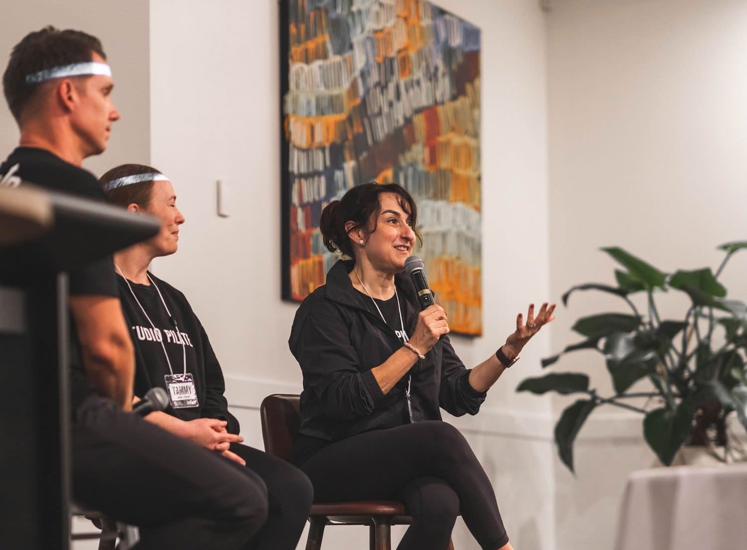 Three seated people indoors, one woman speaking into a microphone, abstract painting and plant in background.