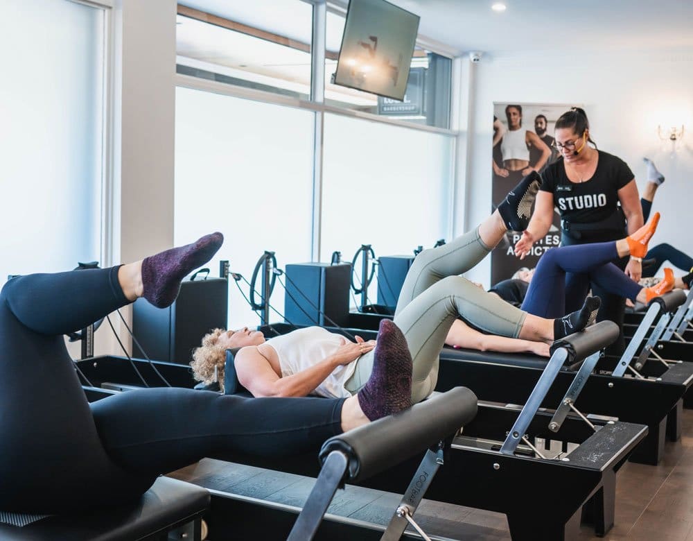 Group Pilates class with participants using reformer machines and an instructor in a studio.