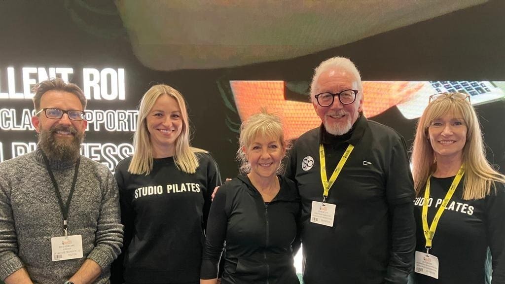 Five people with event badges, two wearing Studio Pilates shirts, standing in front of a screen.