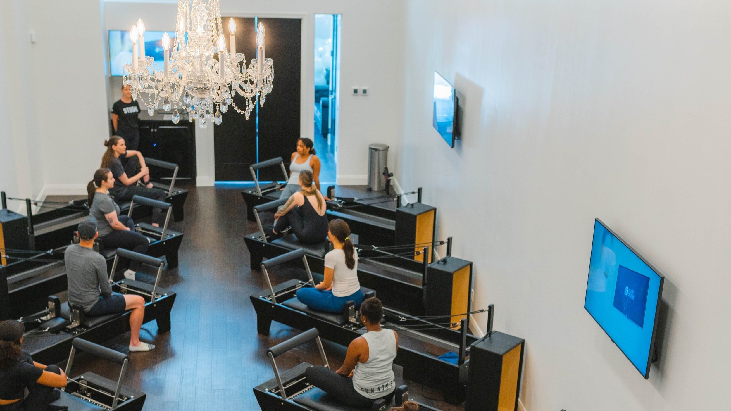 People seated on Pilates reformer machines in a spacious room with chandelier and wall-mounted TVs.
