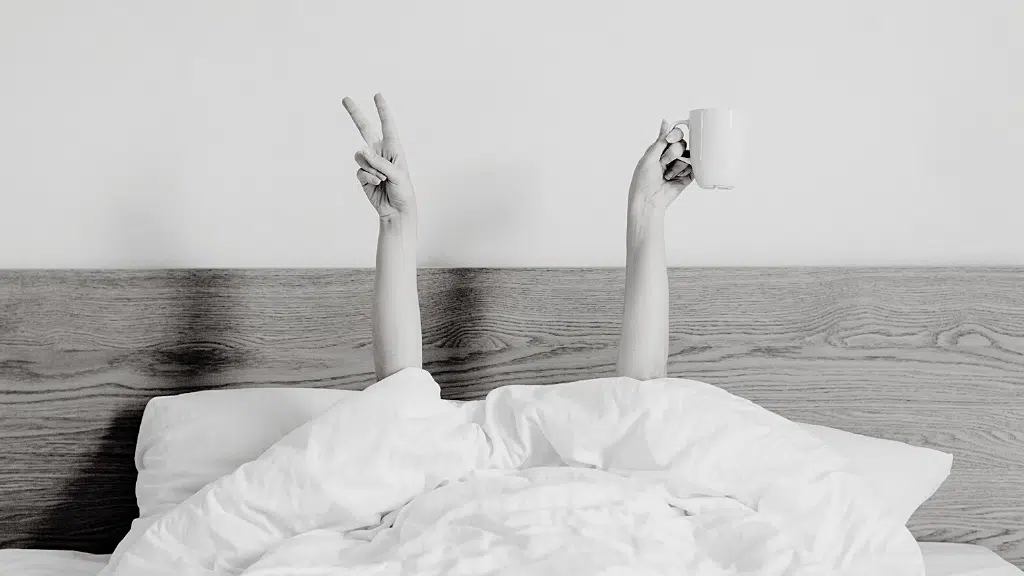 Arms raised from under white blankets on bed, one hand holding a mug, the other showing a peace sign.
