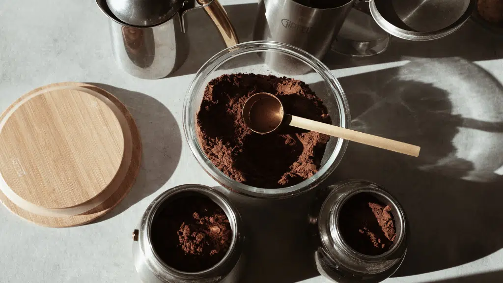 Glass bowl and two metal containers filled with ground coffee, scoop and wooden lid on countertop.