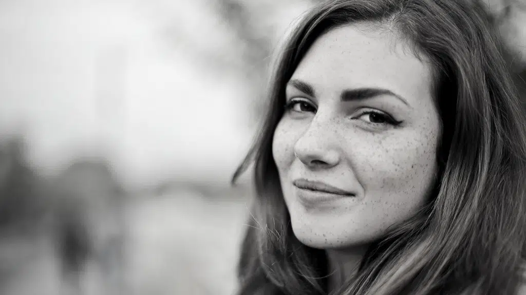 Black and white close-up of a smiling woman with freckles and eyeliner, blurred background.