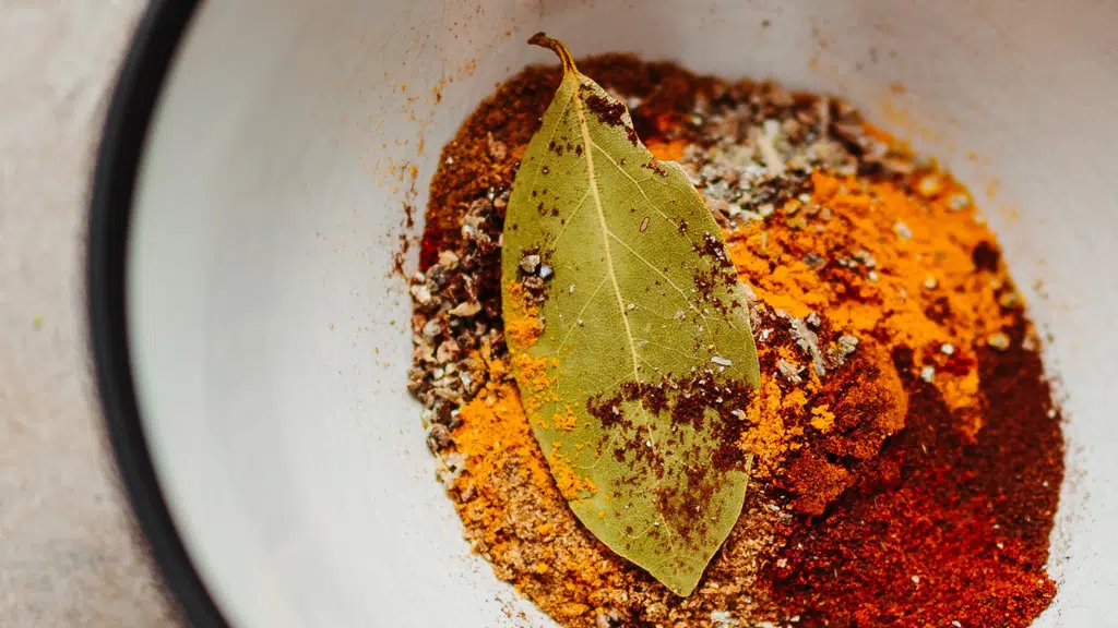 White bowl with ground spices including turmeric, black pepper, and a dried bay leaf on top