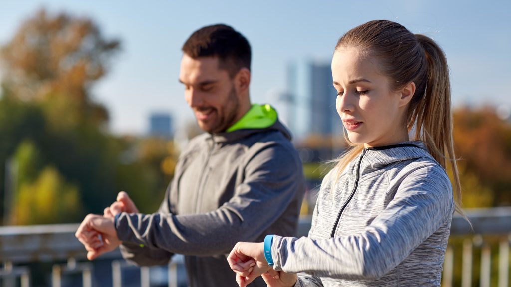 Man and woman in athletic wear outdoors checking their fitness trackers with cityscape background.