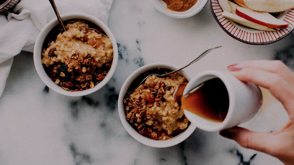 Two bowls of oatmeal topped with granola on marble with syrup being poured into one bowl.