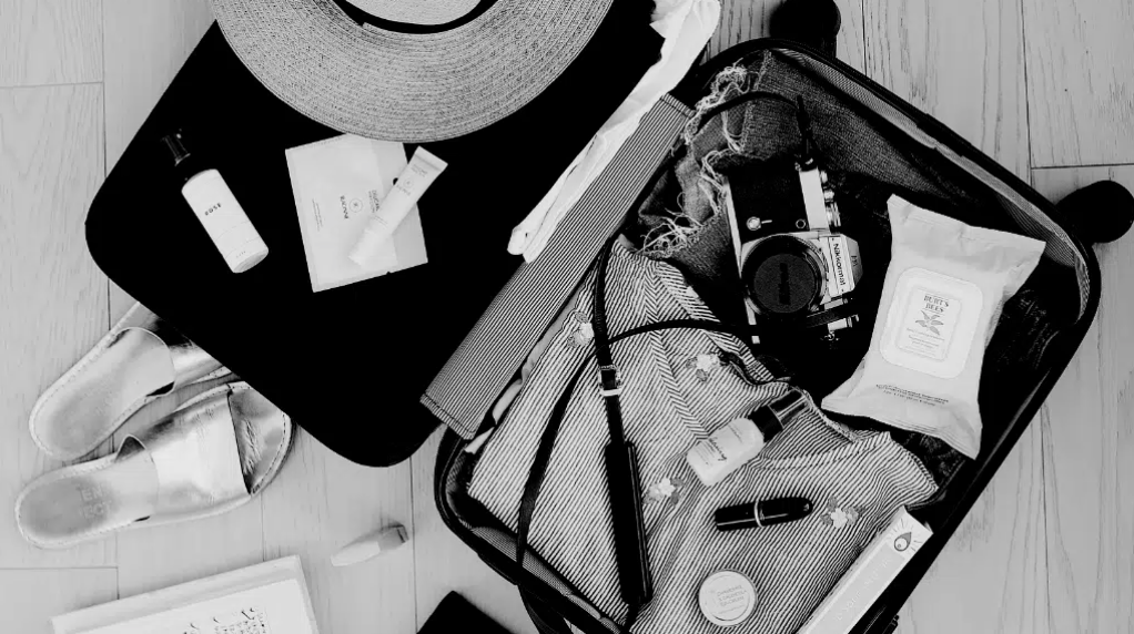 Open suitcase with camera, striped clothing, wipes, lipstick, hat, sandals, and toiletry items on wooden floor.