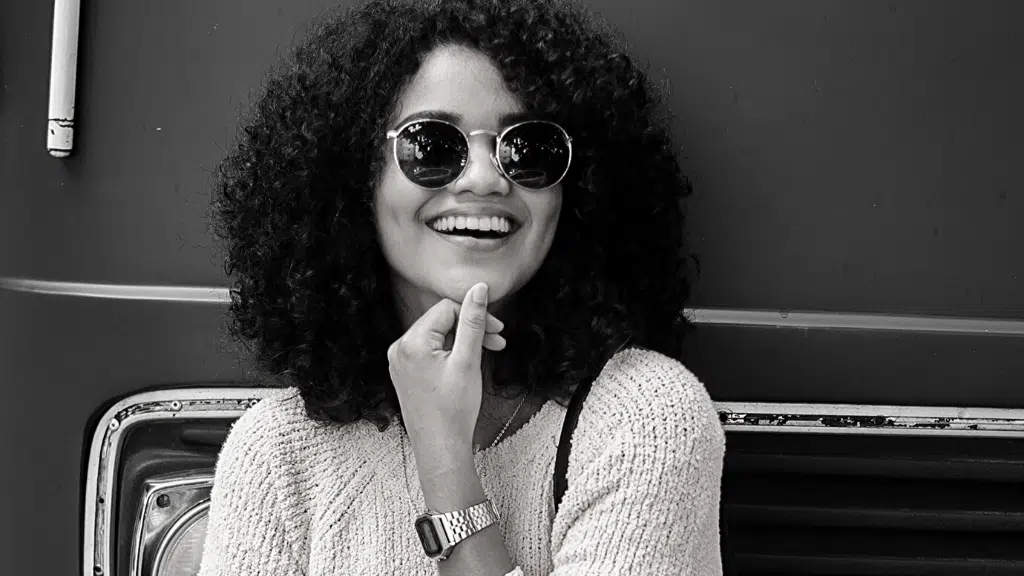 Black and white photo of a smiling woman with curly hair, sunglasses, sweater, and wristwatch.