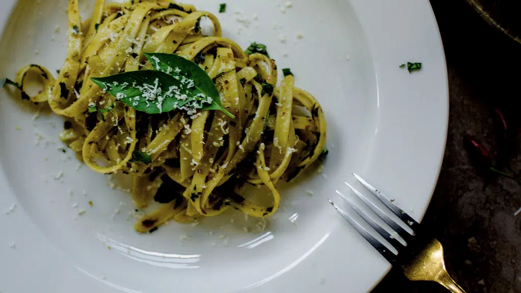 Tagliatelle pasta with grated cheese and basil leaf on a white plate with a fork.