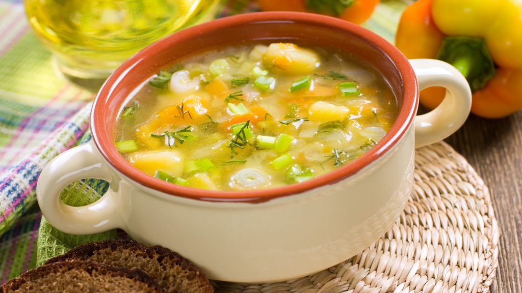 Vegetable soup with carrots, white beans, and herbs in a bowl with bread and yellow pepper background