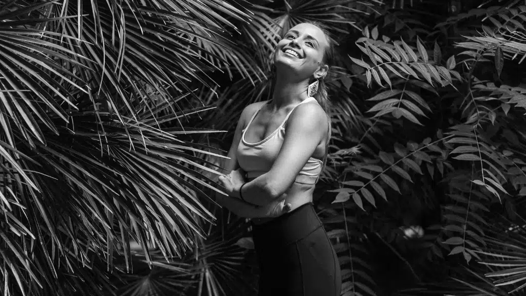 Woman in sportswear standing with arms crossed among dense foliage in black and white.
